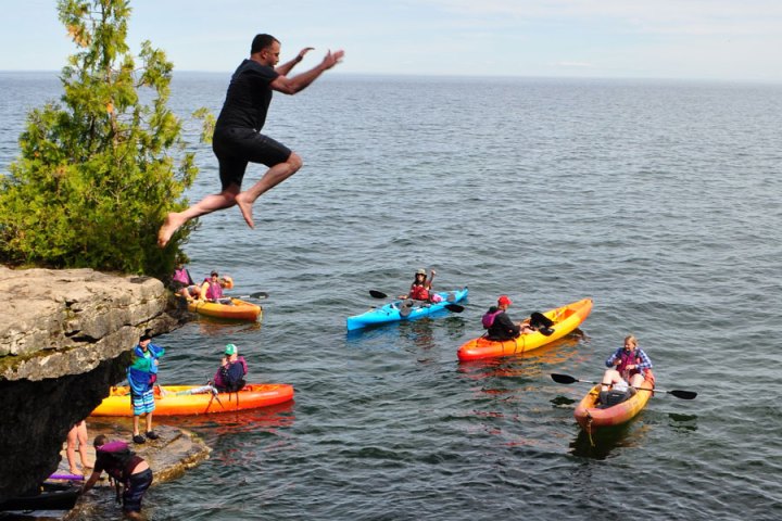 a man in mid air jumping off a cliff at Cave Point County Park with kayaks behind him